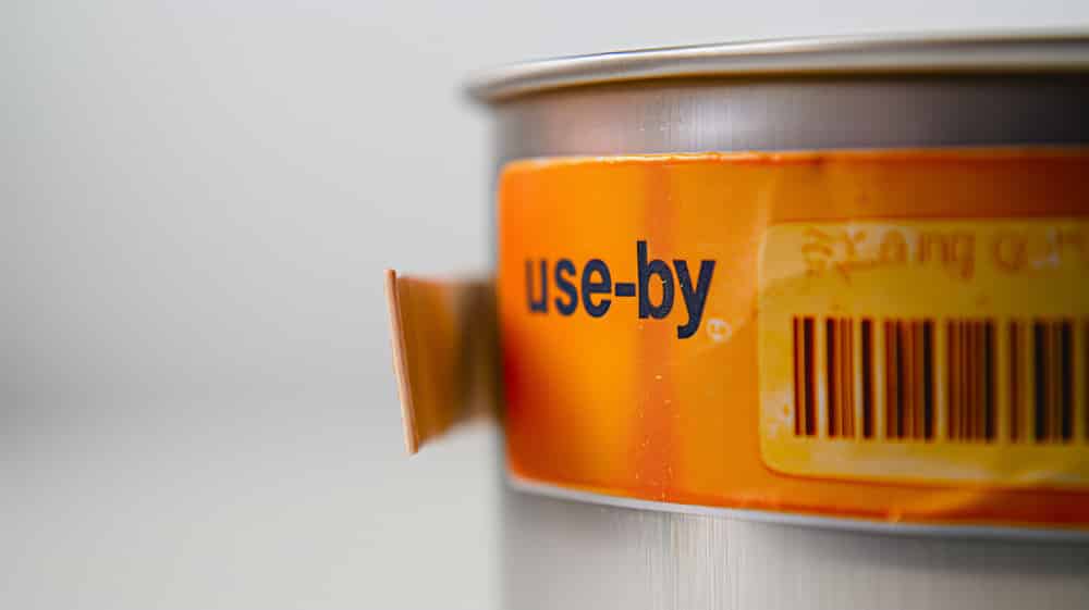 Close-up of an orange "use-by" label on a tin can with a barcode. How to determine freshness and quality?